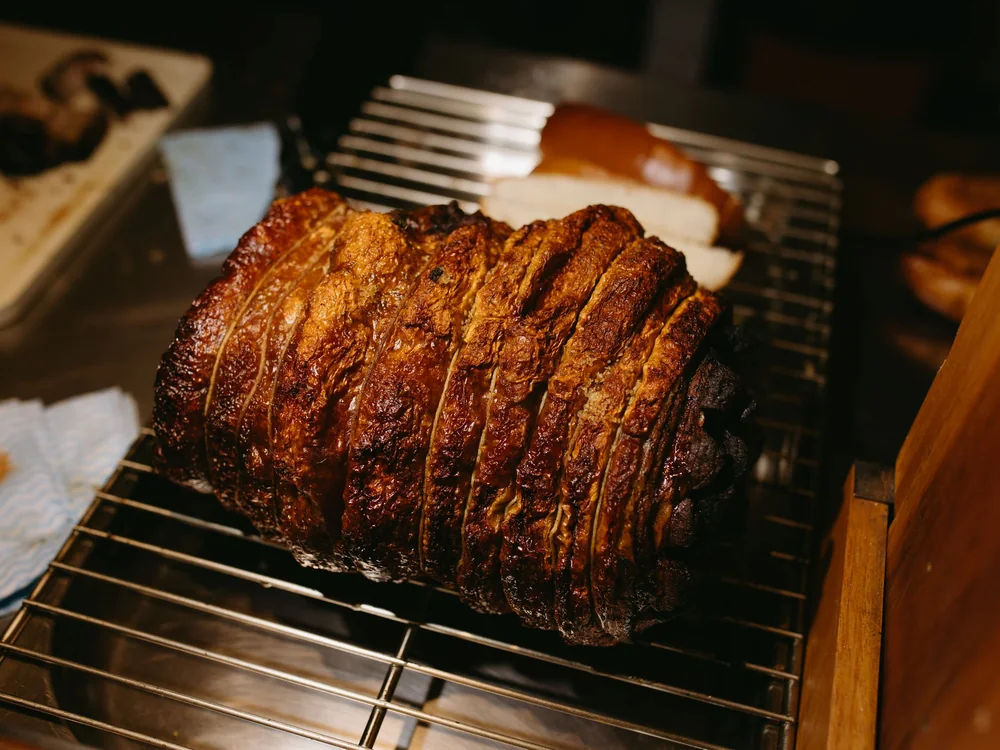 A glazed, sliced roast sitting on a cooling rack beside bread rolls