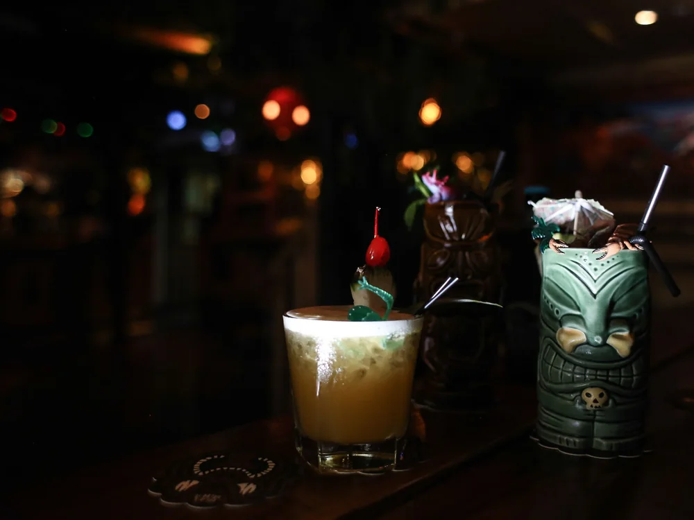 A tropical cocktail with a cherry garnish on a bar counter beside carved tiki mugs